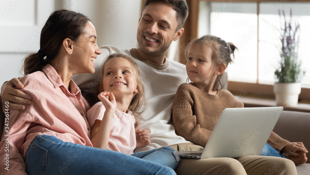 Happy parents with two little daughters using laptop at home, hugging and talking, watching cartoons, shopping online, making video call, family having fun with computer together Happy parents with two little daughters using laptop at home, hugging and talking, watching cartoons, shopping online, making video call, family having fun with computer together