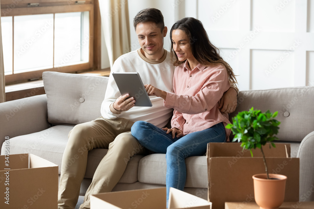 Happy young couple using computer tablet together, looking at mobile device screen, sitting on couch in living room with cardboard boxes with belongings, choosing moving service, relocation concept Happy young couple using computer tablet together, looking at mobile device screen, sitting on couch in living room with cardboard boxes with belongings, choosing moving service, relocation concept