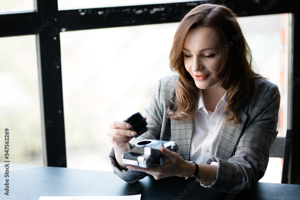 Attractive woman in a plaid jacket holds a manual lens in her hand, unscrewed from the old retro camera, sitting at the table Attractive woman in a plaid jacket holds a manual lens in her hand, unscrewed from the old retro camera, sitting at the table