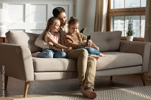 Smiling father with two little daughters using phone, looking at screen, watching cartoons or video, playing game, shopping online, cute preschool girls with dad hugging, sitting on couch at home