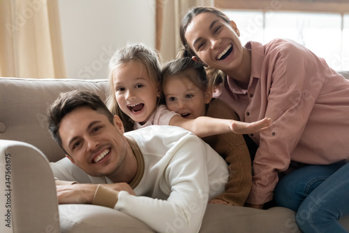 Portrait happy parents with two preschool daughters lying on couch together, looking at camera, smiling mother and father with cute little girls having fun, posing for family photo at home