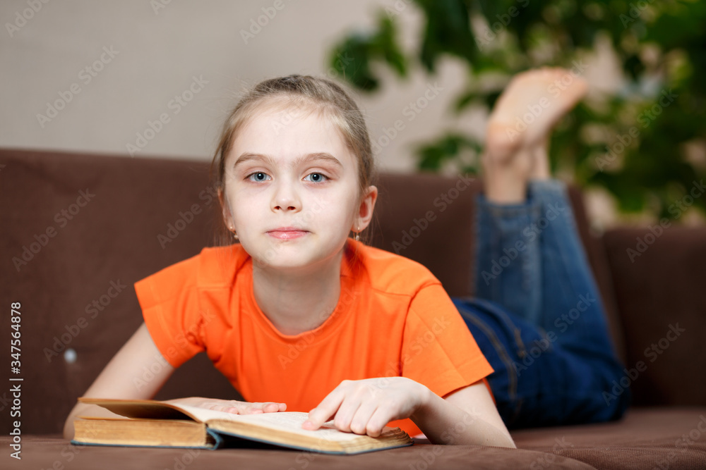 Cute caucasian little girl reading book while lying on sofa at home during quarantine due to coronavirus. Education and leisure concept Cute caucasian little girl reading book while lying on sofa at home during quarantine due to coronavirus. Education and leisure concept