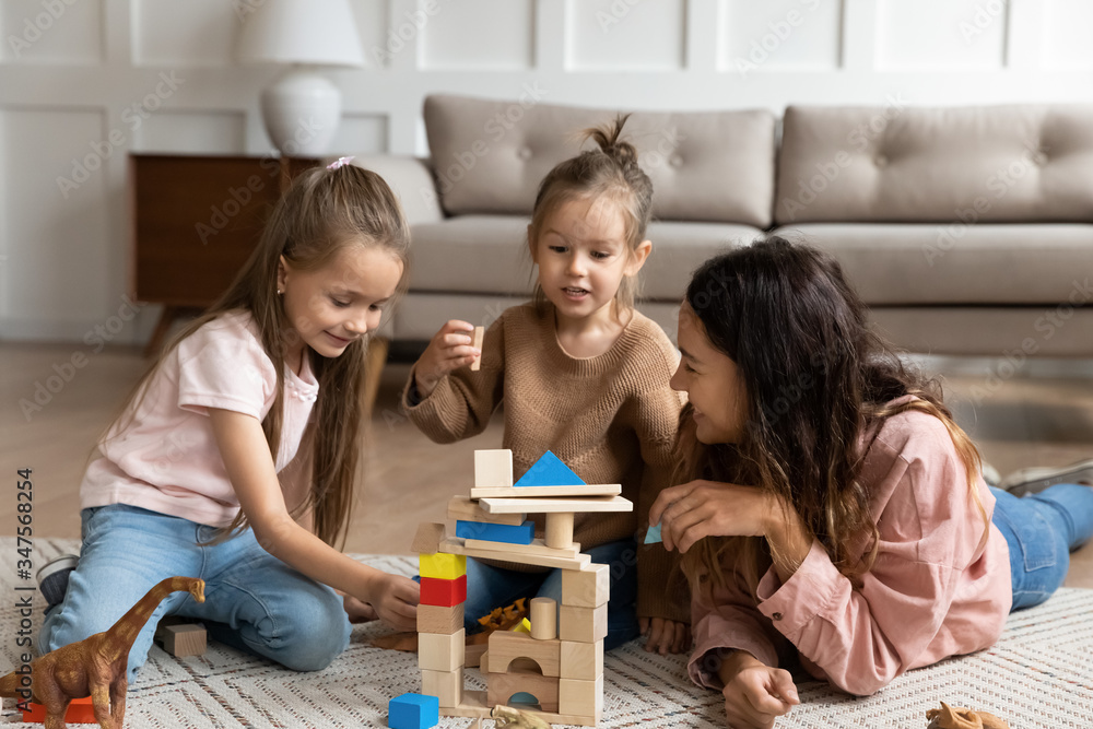 Happy young mother playing with two little daughters, sitting on warm floor in living room, loving mum spending weekend with children at home, cute girls with baby sitter building wooden blocks Happy young mother playing with two little daughters, sitting on warm floor in living room, loving mum spending weekend with children at home, cute girls with baby sitter building wooden blocks