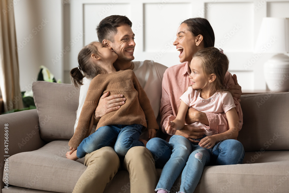 Happy mother and father with little daughters sitting on couch, hugging, two cute preschool girls sitting on smiling parents laps, family spending weekend together, relaxing on cozy sofa