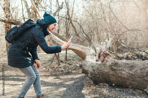 Caucasian woman taking picture photo of squirrel in park. Tourist traveler girl snapping smartphone photos of wild animal in a forest. Fun outdoor activity and blogging vlogging online.