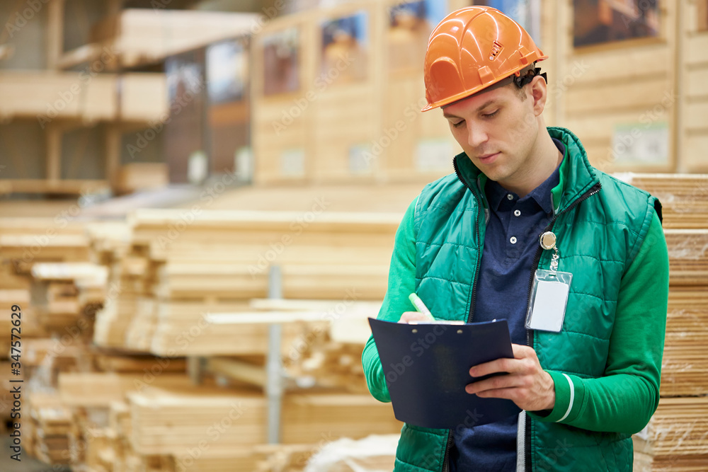 concentrated confident warehouse worker write the number of boards in a document, hard working male work in large distribution center. a lot of wooden boards in the background concentrated confident warehouse worker write the number of boards in a document, hard working male work in large distribution center. a lot of wooden boards in the background