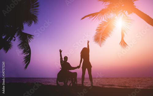 Disabled man in a wheelchair with his wife on the beach. Silhouettes at sunset © romaset