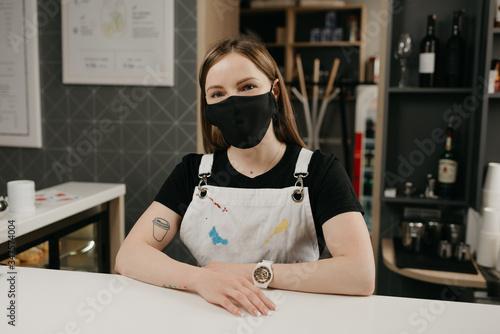 A happy female barista with tattoos in a medical black face mask smiles and waiting for clients in the coffee shop. A beautiful female owner of the cafe poses with arms crossed behind the bar. 