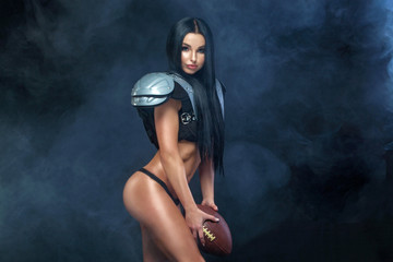 American football. Young sporty brunette wearing sexy uniform of rugby football player posing with ball in smoke isolated on black background