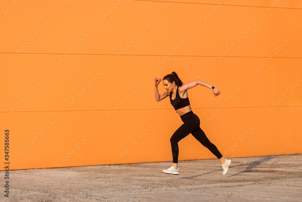 Motivated confident fit woman athlete in tight sportswear, black pants and top, starting to run, jogging outdoor against orange wall, advertising area. Health care and weight loss, sport activity