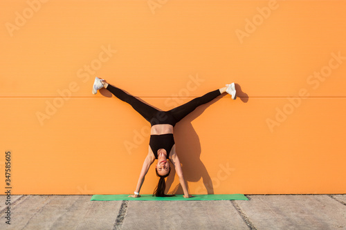 Fototapeta Overjoyed happy girl with perfect athletic body in tight sportswear doing yoga handstand pose with spread legs against wall and showing tongue, having fun