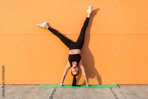 Fototapeta Overjoyed excited girl with perfect athletic body in tight sportswear doing yoga handstand pose against wall and laughing, shouting from happiness