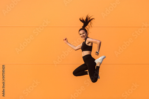 Happy lively vivid girl with fit body in tight sportswear jumping in air, flying and smiling full of energy, healthy sport lifestyle. studio shot, isolated on orange background, advertising area