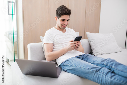 Young handsome man sitting relaxed on sofa with laptop checking his smartphone messages