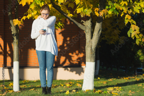 Millennial woman with eyeglasses is frowning at her smartphone while texting and rubbing her shoulder because she is  feeling insecure and anxious. Full length outdoor portrait. 