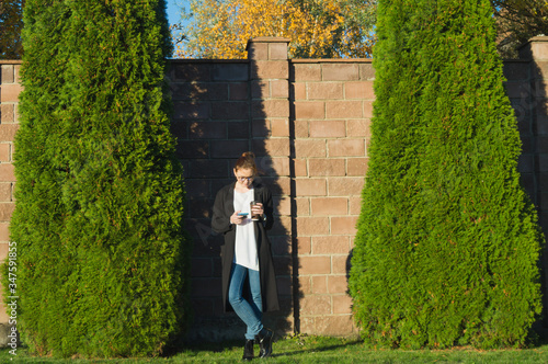 Business woman in park is texting on smartphone, holding cup of coffee. Full length portrait of focused woman in spectacles, black trench coat, white sweater, blue jeans. October evening in Moscow.