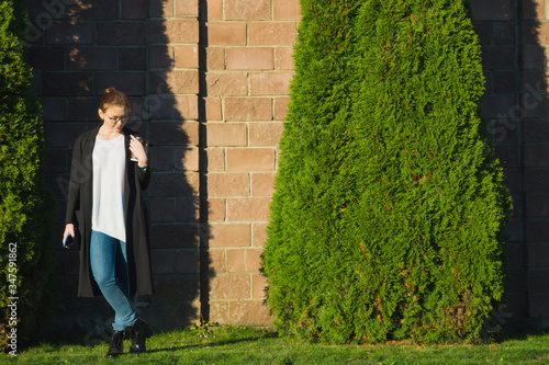Woman standing in thoughtful pose  holding cup of coffee and smartphone. Full length outdoor portrait of woman in spectacles, black trench coat, white sweater, blue jeans. October evening in Moscow.