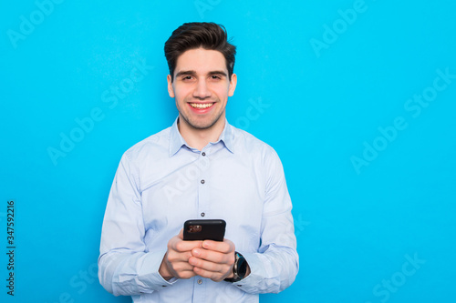 Smiling young business man holding mobile phone, looking at screen isolated on blue studio background. © F8  \ Suport Ukraine