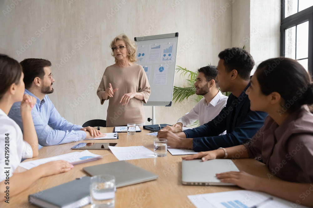 Serious mature 60 year old businesswoman talking, presentation new project in boardroom at company meeting. Adult confident woman coach auditor speaks about business on flip chart graphs background. Serious mature 60 year old businesswoman talking, presentation new project in boardroom at company meeting. Adult confident woman coach auditor speaks about business on flip chart graphs background.