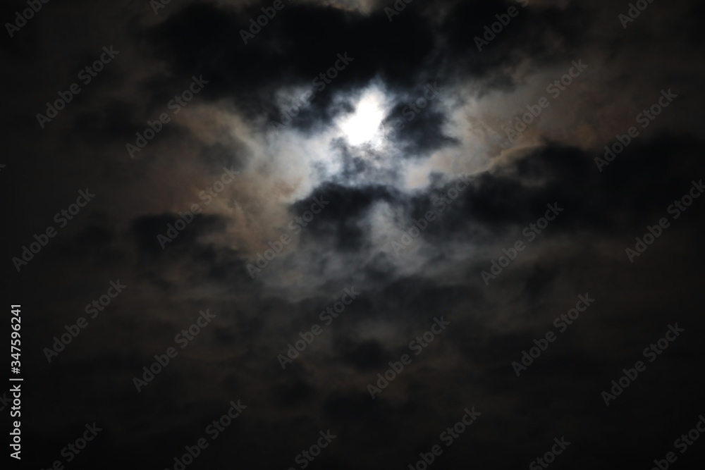 Mystical landscape of the night sky.The outline of the full moon is hidden by black and gray fluffy clouds on a smoky background of voluminous haze that hides the light.A mix of dark tones Mystical landscape of the night sky.The outline of the full moon is hidden by black and gray fluffy clouds on a smoky background of voluminous haze that hides the light.A mix of dark tones