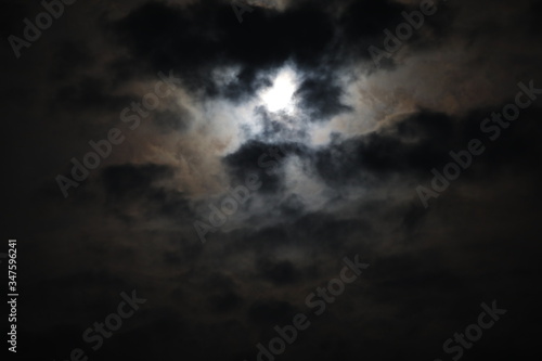 Mystical landscape of the night sky.The outline of the full moon is hidden by black and gray fluffy clouds on a smoky background of voluminous haze that hides the light.A mix of dark tones