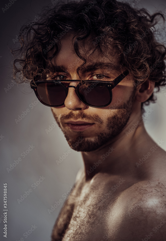 Cool and sexy young man posing in the dark studio shirtless, covering his eyes with a sunglasses and looking into camera Cool and sexy young man posing in the dark studio shirtless, covering his eyes with a sunglasses and looking into camera