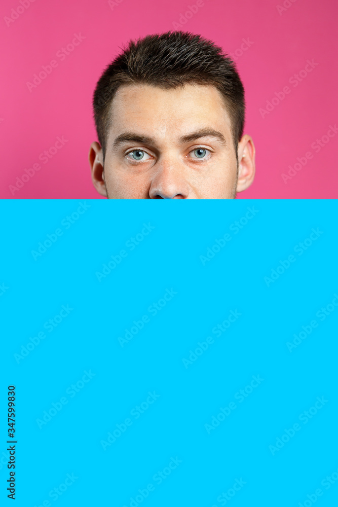 Young handsome man holding a blank cardboard blue banner over an isolated pink background, serious face, thinking about a question. Place for an inscription. Idea for recording thoughts Young handsome man holding a blank cardboard blue banner over an isolated pink background, serious face, thinking about a question. Place for an inscription. Idea for recording thoughts