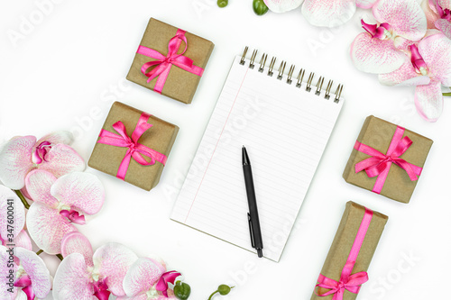 flatlay of Gift present boxes with pink ribbon and orchid flowers on white background top view. Note pad writing.
