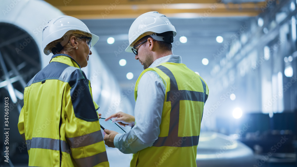 Two Heavy Industry Engineers Stand in Pipe Manufacturing Factory, Use Digital Tablet Computer, Have Discussion. Construction of Oil, Gas and Fuels Transport Pipeline. Back View Sparks Flying Two Heavy Industry Engineers Stand in Pipe Manufacturing Factory, Use Digital Tablet Computer, Have Discussion. Construction of Oil, Gas and Fuels Transport Pipeline. Back View Sparks Flying