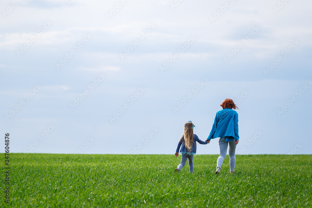 little girl child and mother woman run and jump, green grass in the field, sunny spring weather, smile and joy of the child, blue sky with clouds