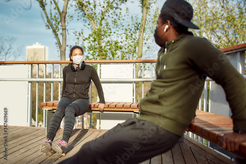 Staying fit during quarantine. Healthy african couple in protective masks working out in park outdoors. Doing push-ups exercises on bench, training triceps photo