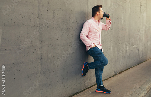 Morning routine. A young stylish man in blue jeans and striped pink shirt drinking coffee or tea leaning against the concrete street wall. City lifestyle. Copy space