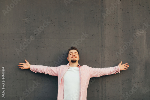 A young happy calm smiling bearded guy with closed eyes and open arms dreaming leaning agains the gray concrete wall with. Copy space