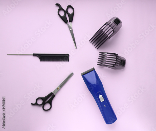 Layout of hairdressing accessories on a lilac background. Hairdressing scissors, comb and a blue hair clipper with two plastic attachments. Selective focus.