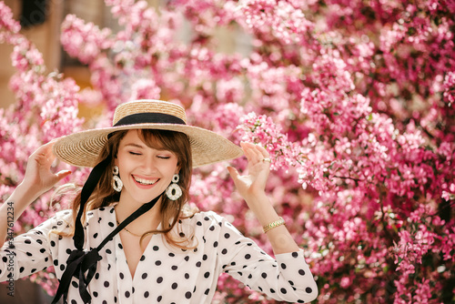 Young beautiful happy smiling girl wearing straw wicker hat, big trendy earrings, polka dot blouse, walking in street, near pink spring blossom trees. Close up portrait. Copy, empty space for text 