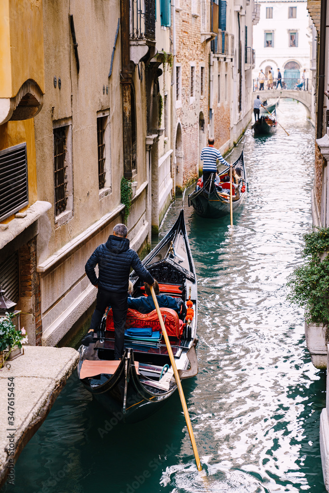 Three gondolas with tourists and gondoliers with paddles and in striped sweaters are sailing along a narrow canal between houses in Venice, Italy. Classic entertainment and excursion in the city