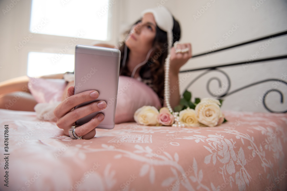 Beautiful young woman holding smart phone while laying in bed Beautiful young woman holding smart phone while laying in bed