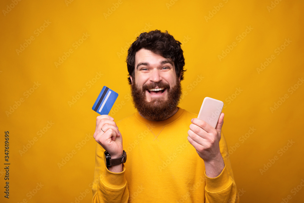 Smiling bearded hipster is holding a phone and a credit or debit card on yellow background. Smiling bearded hipster is holding a phone and a credit or debit card on yellow background.
