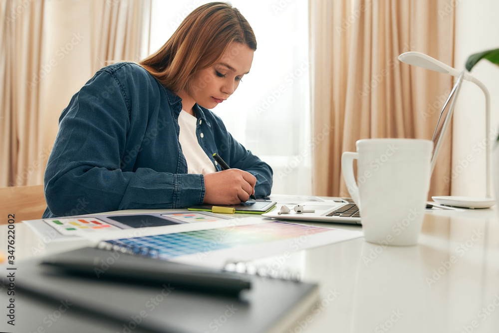 Female designer working from home. Young creative woman in casual clothes drawing something on graphic tablet while sitting at her creative workplace at home Female designer working from home. Young creative woman in casual clothes drawing something on graphic tablet while sitting at her creative workplace at home
