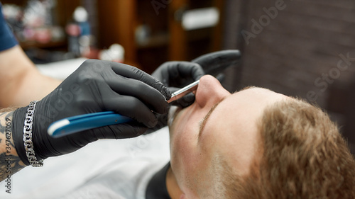 Dangerous razor. Male barber in black gloves with tattooed arm shaving client with straight razor. Traditional shave. Web banner