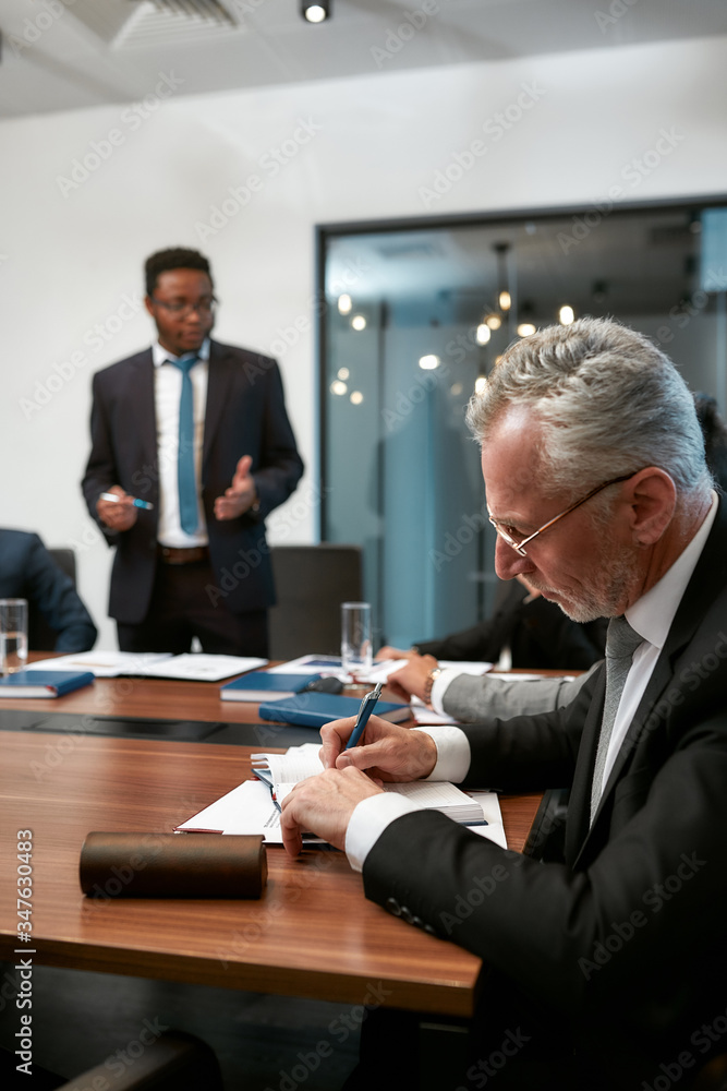 Focused mature caucasian businessman writing something in his note pad while having a meeting in the modern office Focused mature caucasian businessman writing something in his note pad while having a meeting in the modern office