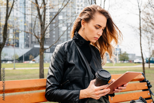 Young woman sitting on the park bench and reading book with tablet, holding the take away coffee, lifestyle concept