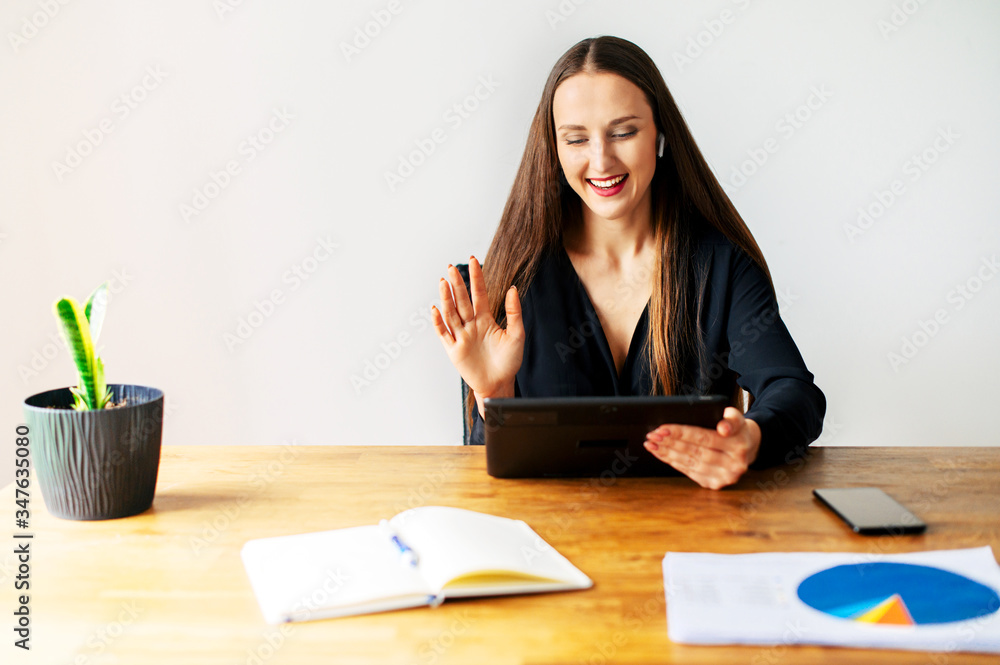 Virtual meeting, video call. A young attractive woman is using tablet for online communicating on the distance. She sits at the table in the office with tablet in hand and waving hello into webcam Virtual meeting, video call. A young attractive woman is using tablet for online communicating on the distance. She sits at the table in the office with tablet in hand and waving hello into webcam