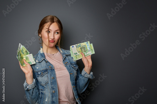 Portrait of a natural and authentic beautiful young woman, teenager with many 100 Euro banknotes in her hand is happy about the money, in front of a grey wall
