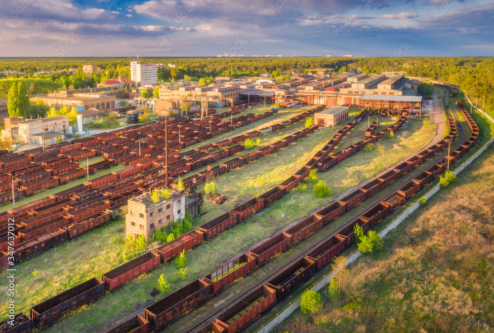 Aerial view of freight trains. Railway station. Rusty wagons on railroad. Heavy industry. Industrial landscape with train in depot, green trees, buildings, sky at sunset. Top view. Transportation