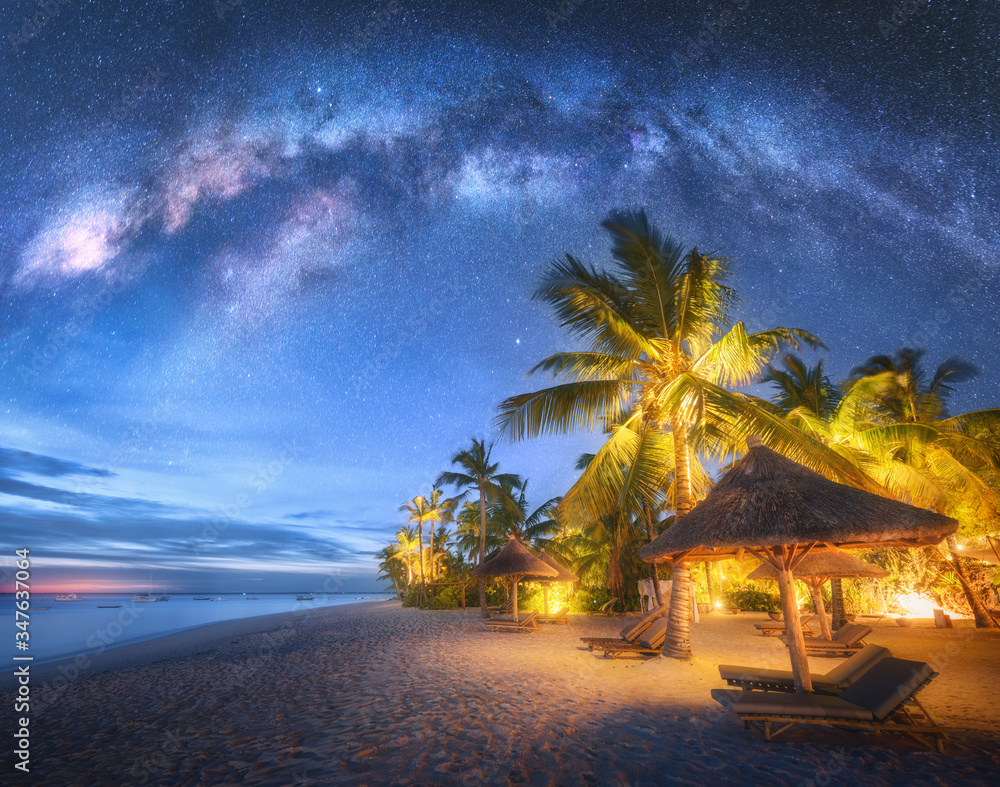 Milky Way over the sandy beach with palm trees and sunbeds and umbrellas at night in summer. Landscape with sea coast, beautiful blue starry sky, galaxy, green palms. Travel in Zanzibar, Africa. Space - obrazy, fototapety, plakaty 