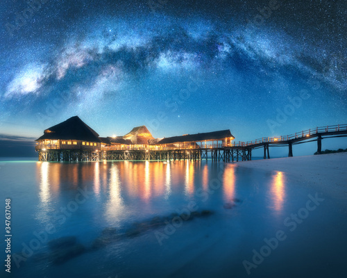 Milky Way over wooden bungalow on the water in summer starry night. Landscape with hotel on the sea, illumination, jetty, sandy beach, sky with stars, reflection in water in Zanzibar, Africa. Space