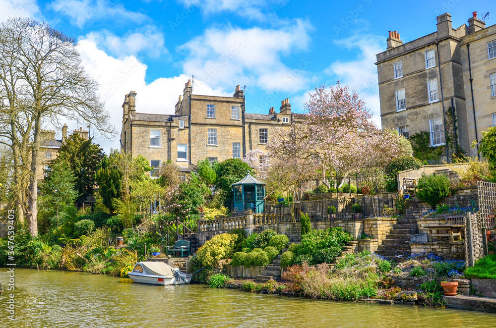 Fototapeta premium Historic houses and beautiful gardens with green plants and flowers by Canal river on a sunny spring day. Boat parked on the heritage waterway - Kennet and Avon Canal, Bath/United Kingdom.