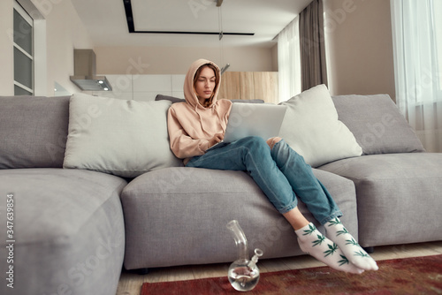 Experience the difference. Young caucasian woman sitting on the couch at home, trying to use laptop after smoking marijuana from a bong or glass water pipe