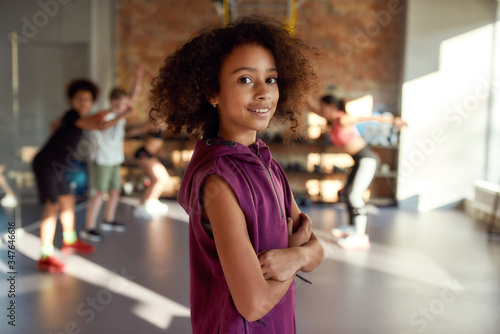 Active kid. Portrait of a girl smiling at camera before warming up, exercising together with other kids and trainer in gym. Sport, healthy lifestyle, active childhood concept